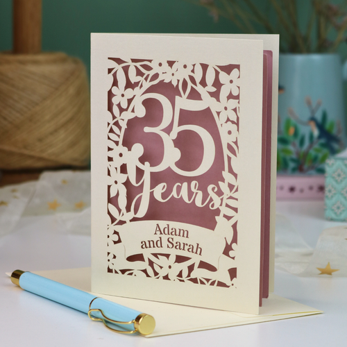 A personalized coral anniversary card with laser-cut floral details and the text '35 Years Adam and Sarah', displayed on a table with a light blue pen in front. The background features a soft-focus teal wall, a vase of dried flowers, and a spool of thread.
