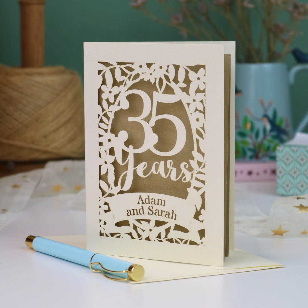 A personalized 35th anniversary card with laser-cut floral details and the text '35 Years Adam and Sarah', displayed on a table with a light blue pen in front. The background features a soft-focus teal wall, a vase of dried flowers, and a spool of thread.
