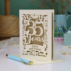 A personalized 35th anniversary card with laser-cut floral details and the text '35 Years Adam and Sarah', displayed on a table with a light blue pen in front. The background features a soft-focus teal wall, a vase of dried flowers, and a spool of thread.