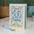 A personalized 35th anniversary card with laser-cut floral details and the text '35 Years Adam and Sarah', displayed on a table with a light blue pen in front. The background features a soft-focus teal wall, a vase of dried flowers, and a spool of thread.