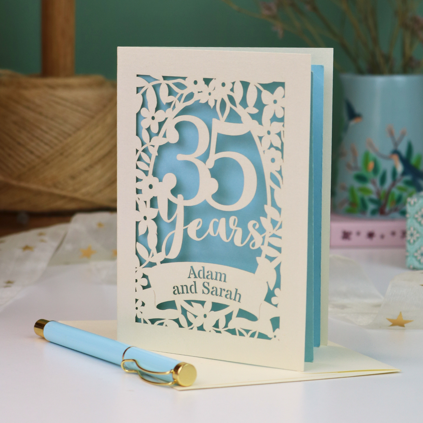 A personalized 35th anniversary card with laser-cut floral details and the text '35 Years Adam and Sarah', displayed on a table with a light blue pen in front. The background features a soft-focus teal wall, a vase of dried flowers, and a spool of thread.