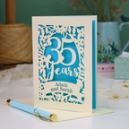 A personalized laser cut 35th anniversary card with laser-cut floral details and the text '35 Years Adam and Sarah', displayed on a table with a light blue pen in front. The background features a soft-focus teal wall, a vase of dried flowers, and a spool of thread.
