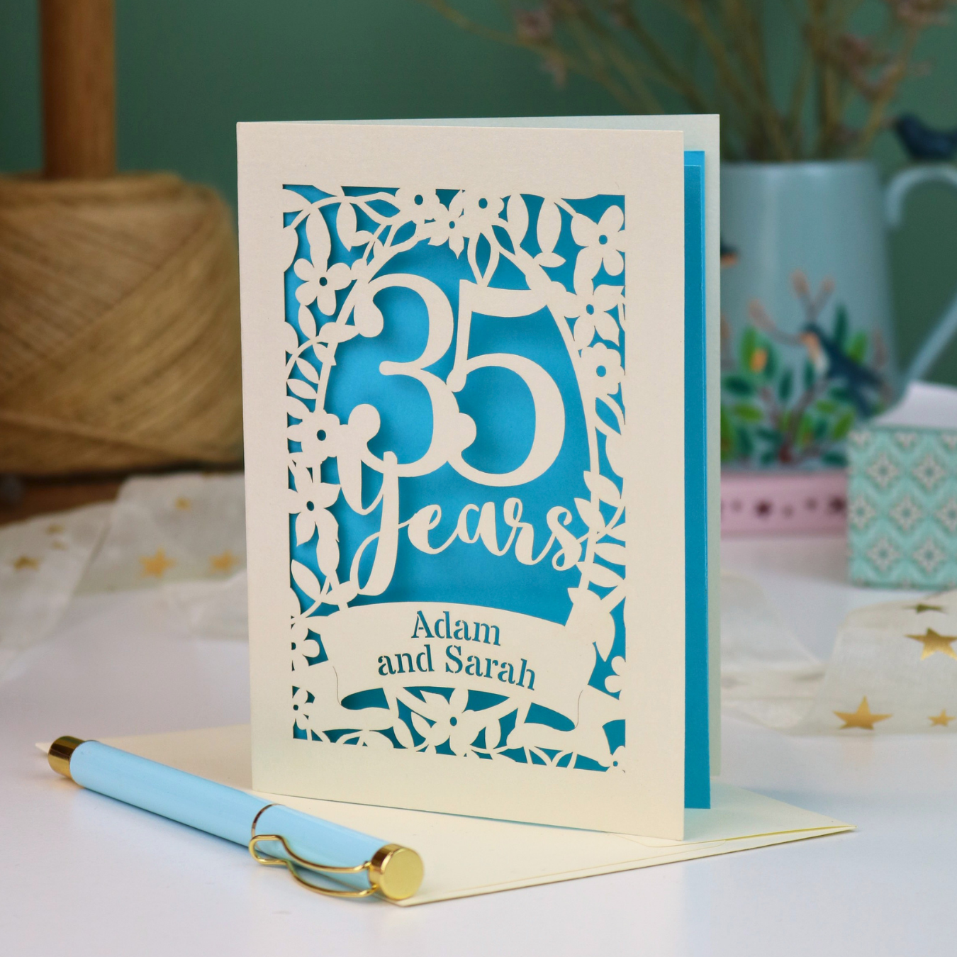 A personalized laser cut 35th anniversary card with laser-cut floral details and the text '35 Years Adam and Sarah', displayed on a table with a light blue pen in front. The background features a soft-focus teal wall, a vase of dried flowers, and a spool of thread.