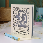 A personalized 35th wedding anniversary card with laser-cut floral details and the text '35 Years Adam and Sarah', displayed on a table with a light blue pen in front. The background features a soft-focus teal wall, a vase of dried flowers, and a spool of thread.