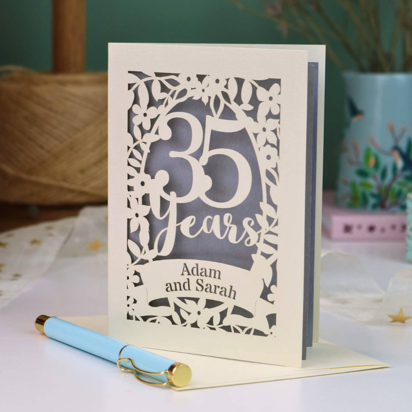 A personalized 35th wedding anniversary card with laser-cut floral details and the text '35 Years Adam and Sarah', displayed on a table with a light blue pen in front. The background features a soft-focus teal wall, a vase of dried flowers, and a spool of thread.