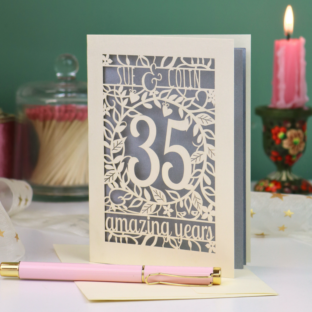 A 35th wedding anniversary laser-cut greeting card with a cream and silver design, displayed on a white surface with a pink pen in the foreground and decorative candles in the background.