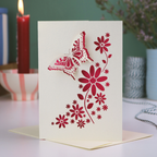 A laser cut Mother's Day card with 3D butterfly and flower design, on a white desk with green background and candle. 