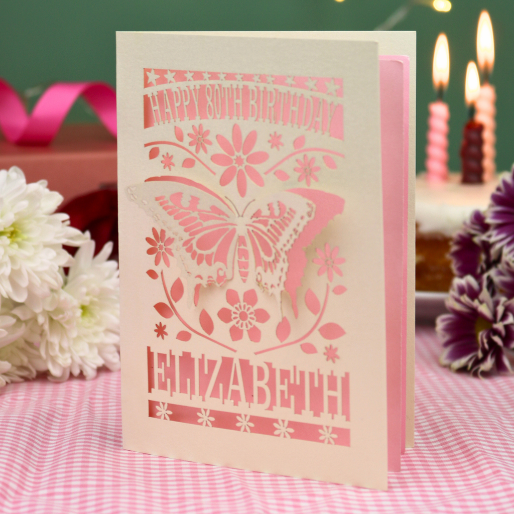 A 3D papercut 80th birthday card for Elizabeth, featuring a butterfly and floral design on a cream card, standing on a pink gingham tablecloth with candles and flowers in the background.