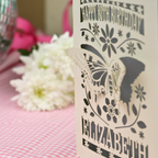 A 3D papercut 80th birthday card for Elizabeth, featuring a butterfly and floral design on a cream card, standing on a pink gingham tablecloth with candles and flowers in the background.