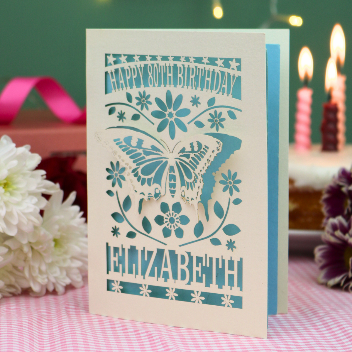 A laser-cut 80th birthday card with a 3D butterfly and floral design, standing on a pink checked tablecloth with blurred birthday candles and flowers in the background.