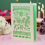 A 3D papercut 80th birthday card for Elizabeth, featuring a butterfly and floral design on a cream card, standing on a pink gingham tablecloth with candles and flowers in the background.
