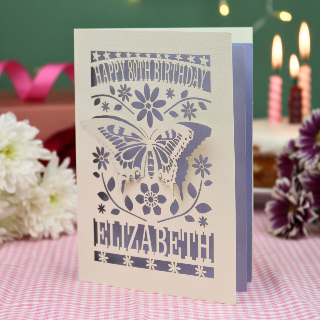A 3D papercut 80th birthday card for Elizabeth, featuring a butterfly and floral design on a cream card, standing on a pink gingham tablecloth with candles and flowers in the background.
