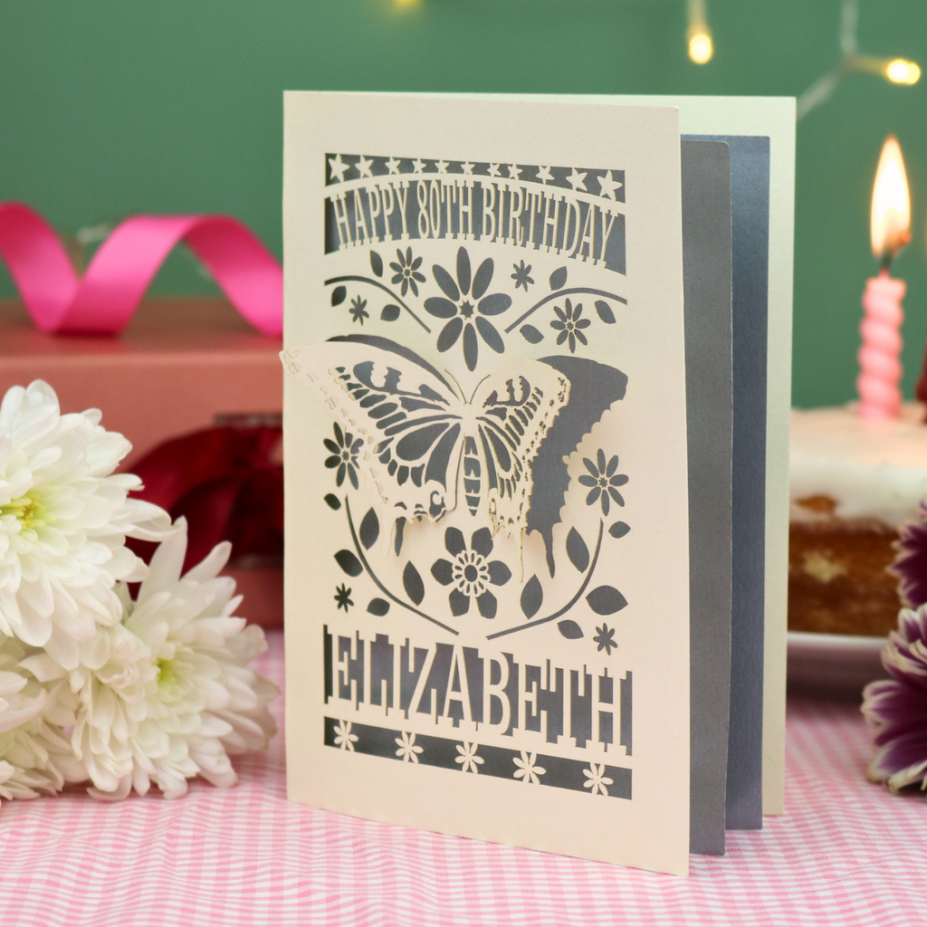 A 3D papercut 80th birthday card for Elizabeth, featuring a butterfly and floral design on a cream card, standing on a pink gingham tablecloth with candles and flowers in the background.