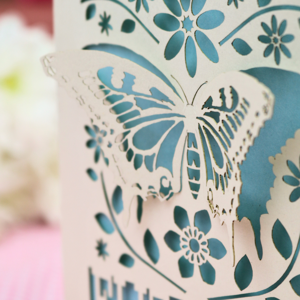 A laser-cut 80th birthday card with a 3D butterfly and floral design, standing on a pink checked tablecloth with blurred birthday candles and flowers in the background.