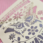 A laser-cut 80th birthday card with a 3D butterfly and floral design, standing on a pink checked tablecloth with blurred birthday candles and flowers in the background.