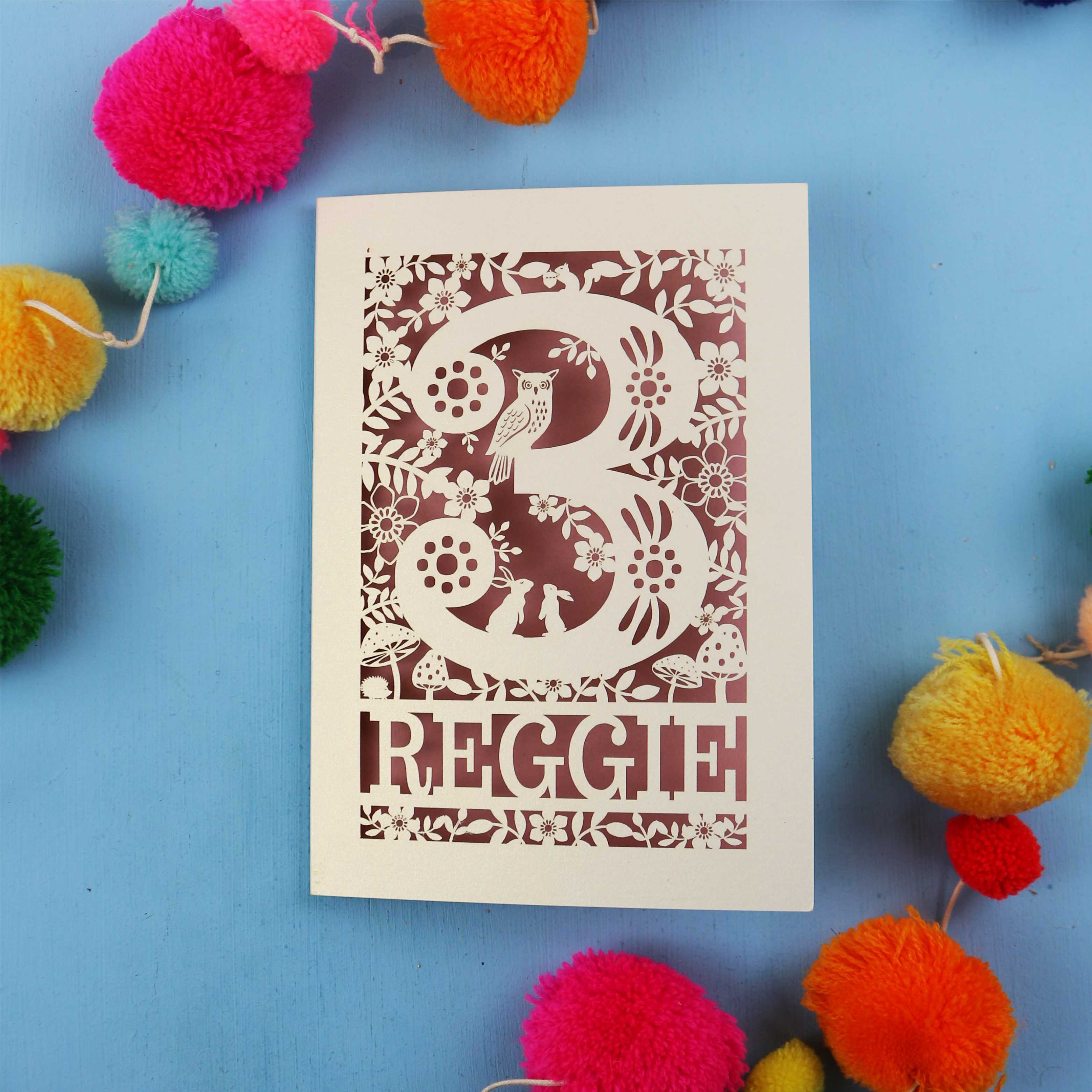 Personalised paper cut 3rd birthday card with floral patterns and 'REGGIE' text on a blue background with colourful pom-poms.