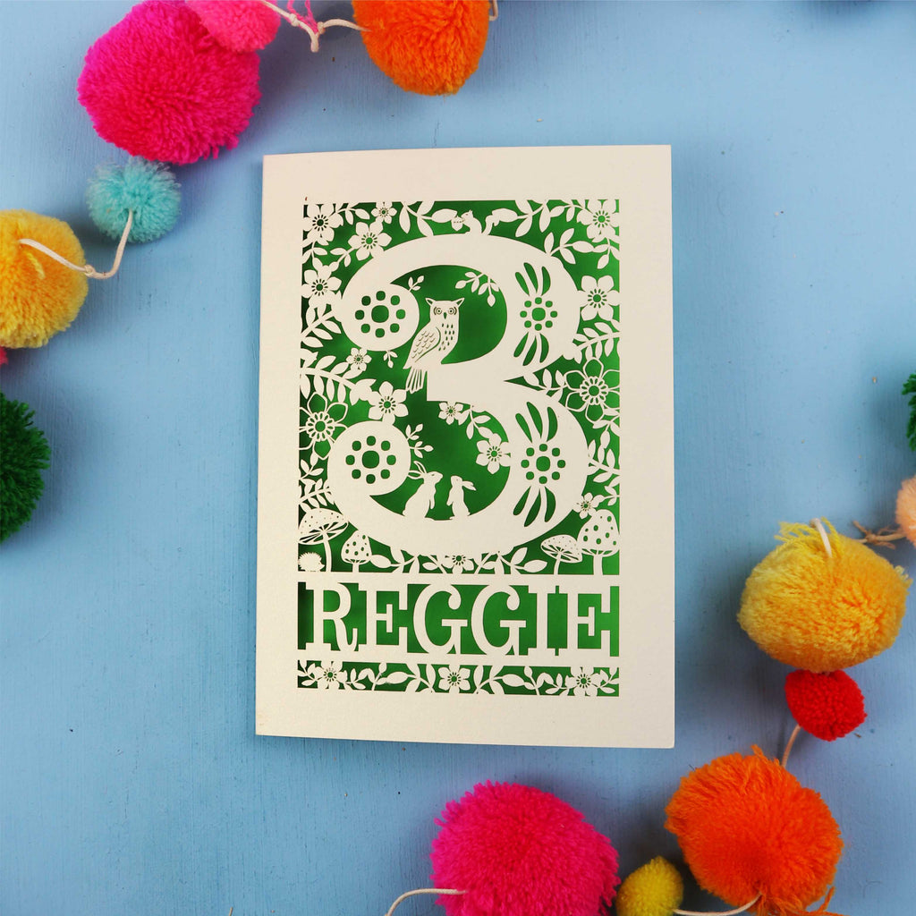 Personalised paper cut third birthday card with floral patterns and 'REGGIE' text on a blue background with colourful pom-poms.