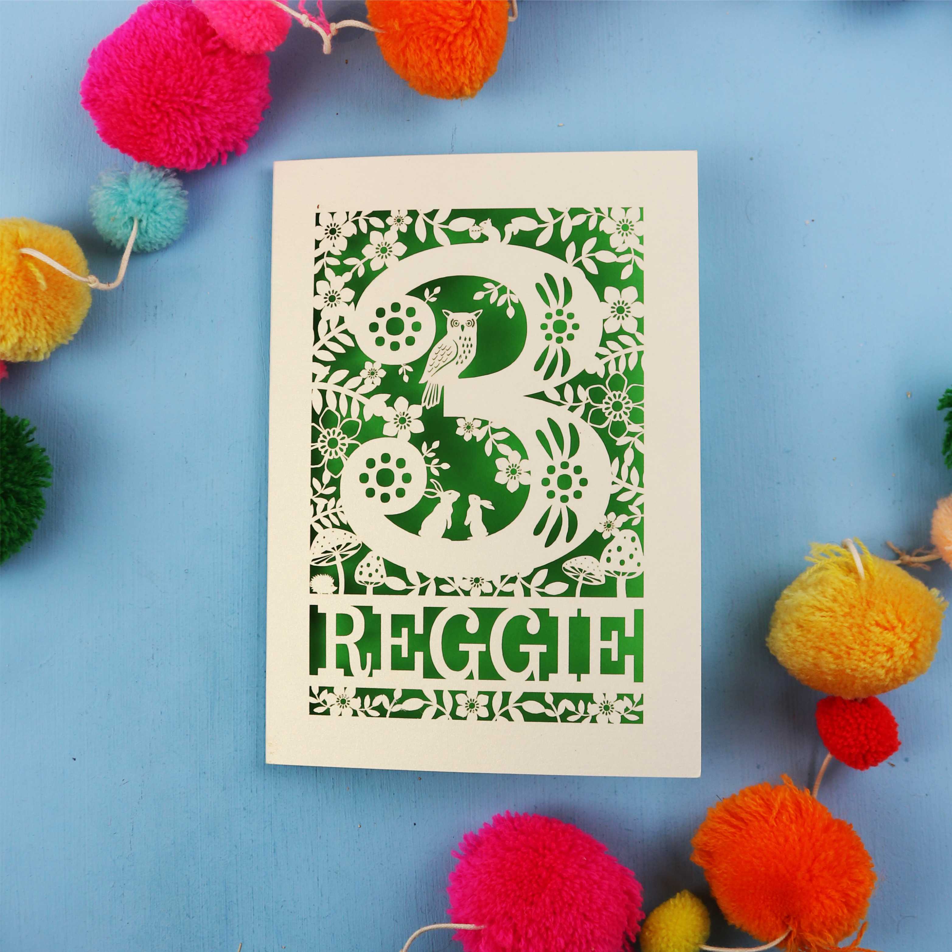 Personalised paper cut third birthday card with floral patterns and 'REGGIE' text on a blue background with colourful pom-poms.