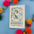 Personalised laser cut 3rd birthday card with floral patterns and 'REGGIE' text on a blue background with colourful pom-poms.