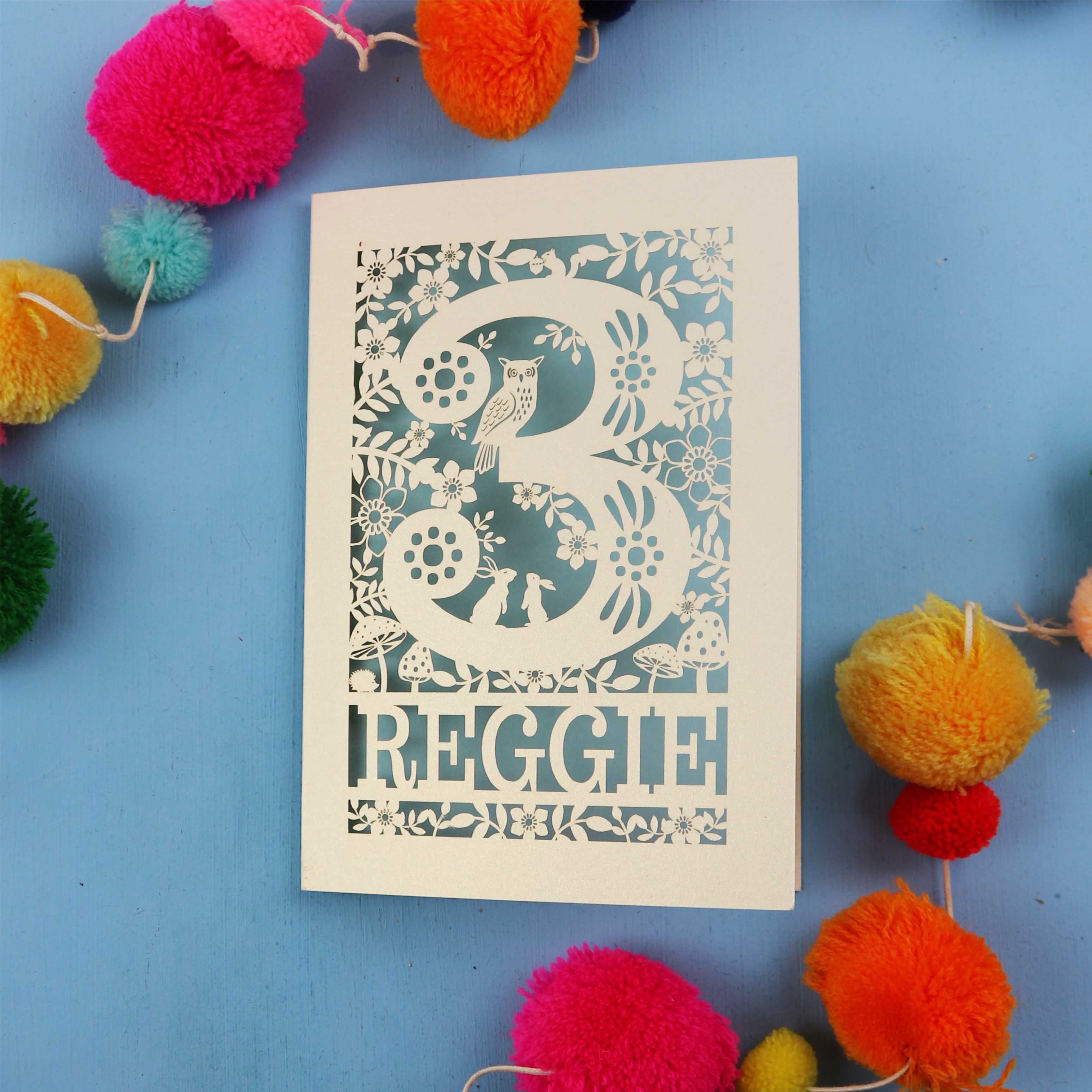 Personalised laser cut 3rd birthday card with floral patterns and 'REGGIE' text on a blue background with colourful pom-poms.