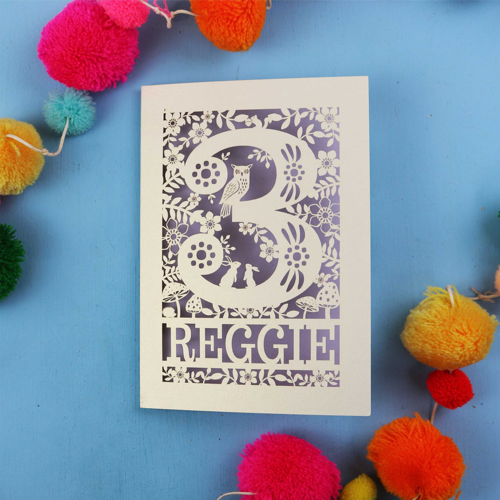 Personalised 3rd birthday card with floral patterns and 'REGGIE' text on a blue background with colourful pom-poms.