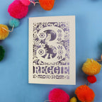 Personalised 3rd birthday card with floral patterns and 'REGGIE' text on a blue background with colourful pom-poms.
