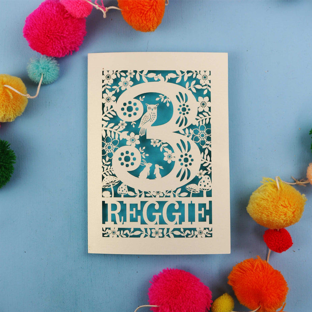 Personalised third birthday card with floral patterns and 'REGGIE' text on a blue background with colourful pom-poms.