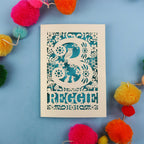 Personalised third birthday card with floral patterns and 'REGGIE' text on a blue background with colourful pom-poms.