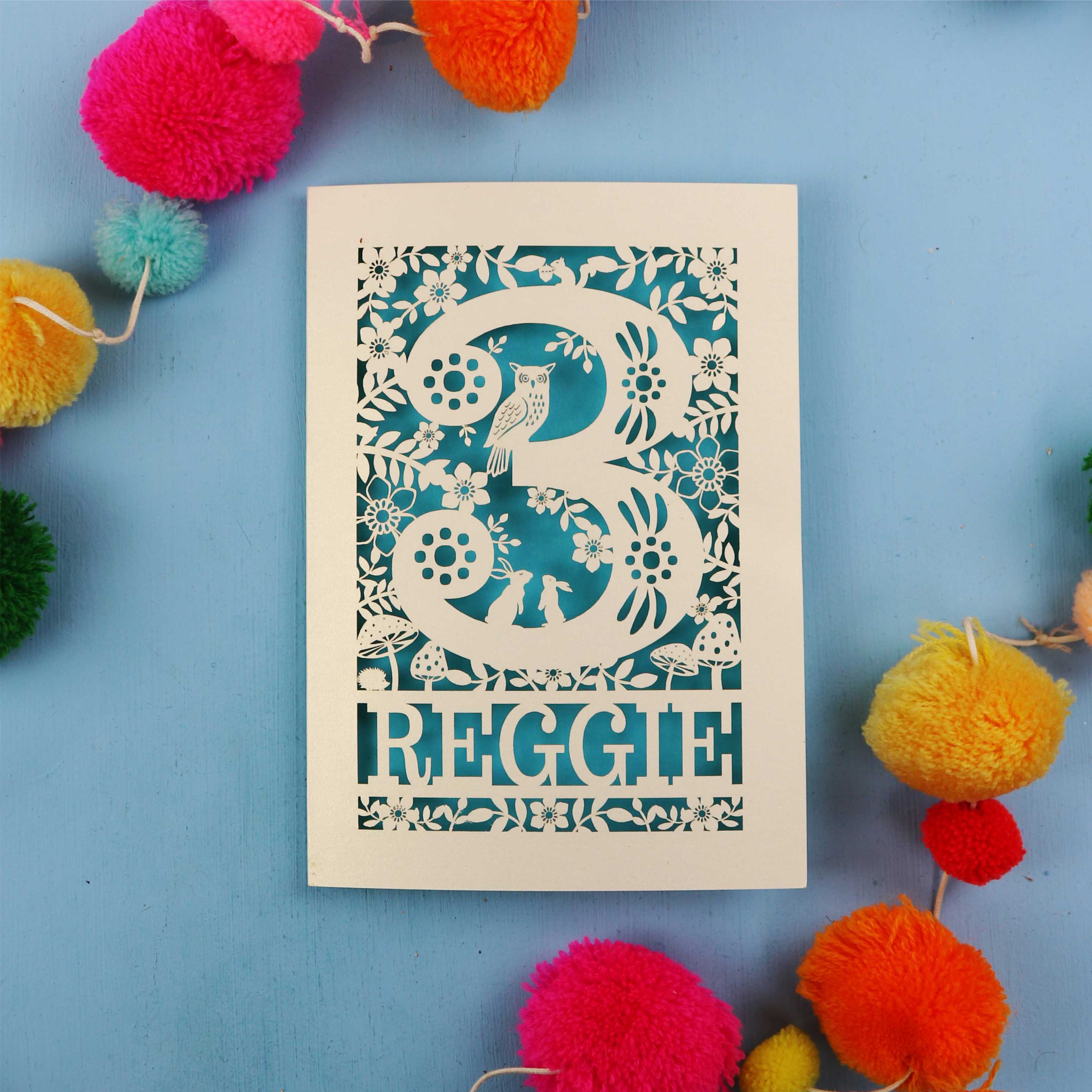 Personalised third birthday card with floral patterns and 'REGGIE' text on a blue background with colourful pom-poms.