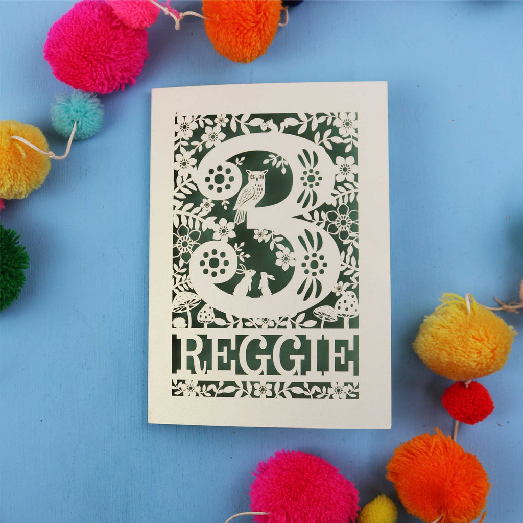 Decorative paper cut out 3rd birthday card with floral patterns and 'REGGIE' text on a blue background with colourful pom-poms.