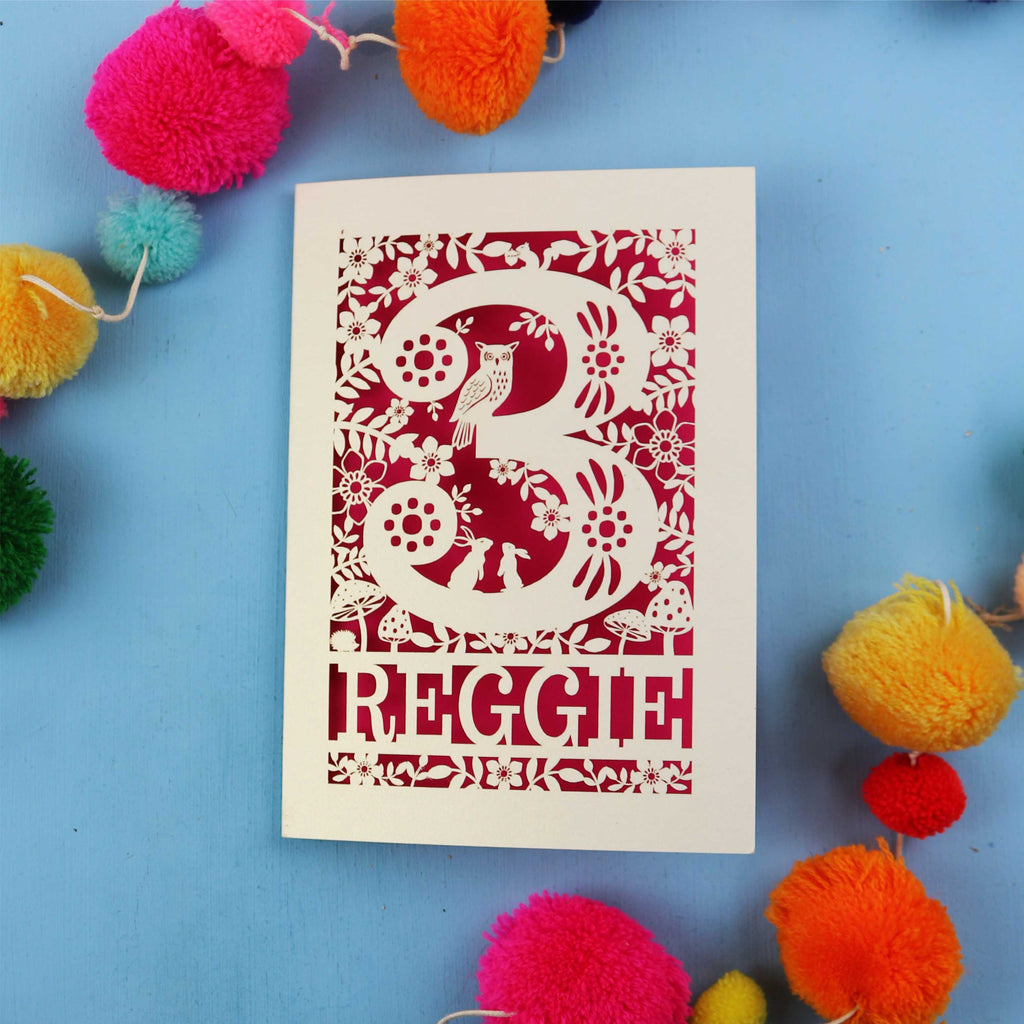Personalised 3rd birthday card with floral patterns and 'REGGIE' text on a blue background with colourful pom-poms.