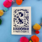 Personalised paper cut 3rd birthday card with floral patterns and 'REGGIE' text on a blue background with colourful pom-poms.