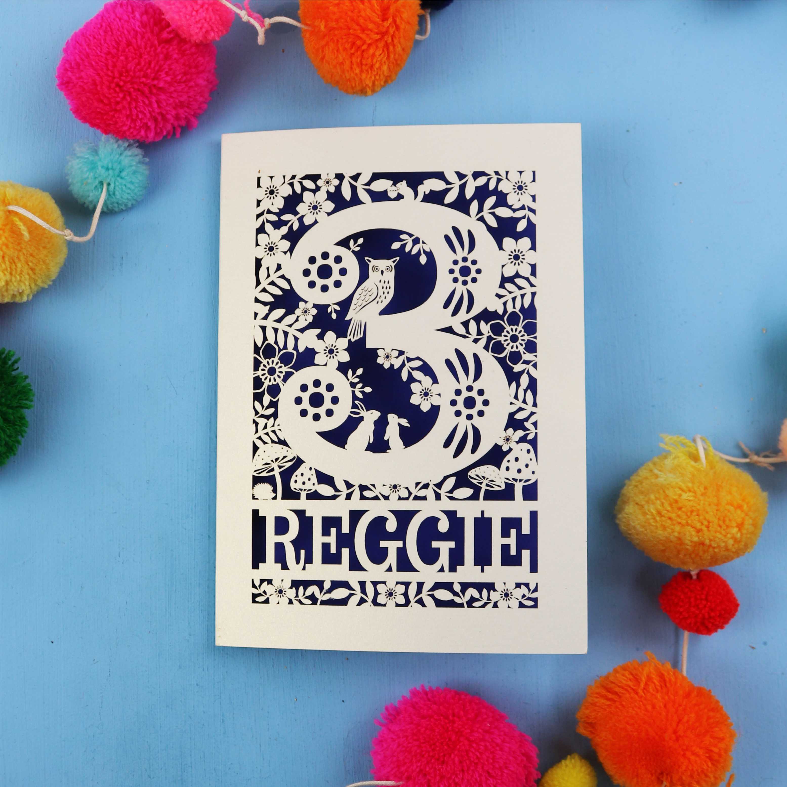 Personalised paper cut 3rd birthday card with floral patterns and 'REGGIE' text on a blue background with colourful pom-poms.