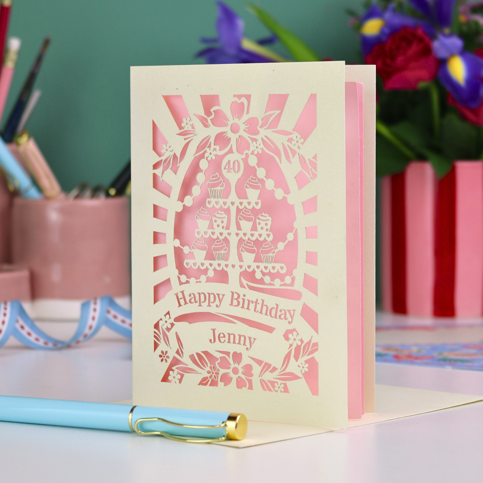 Personalized laser cut 40th birthday card with decorative cut-out cupcakes on a cake stand design on a desk with stationery items.