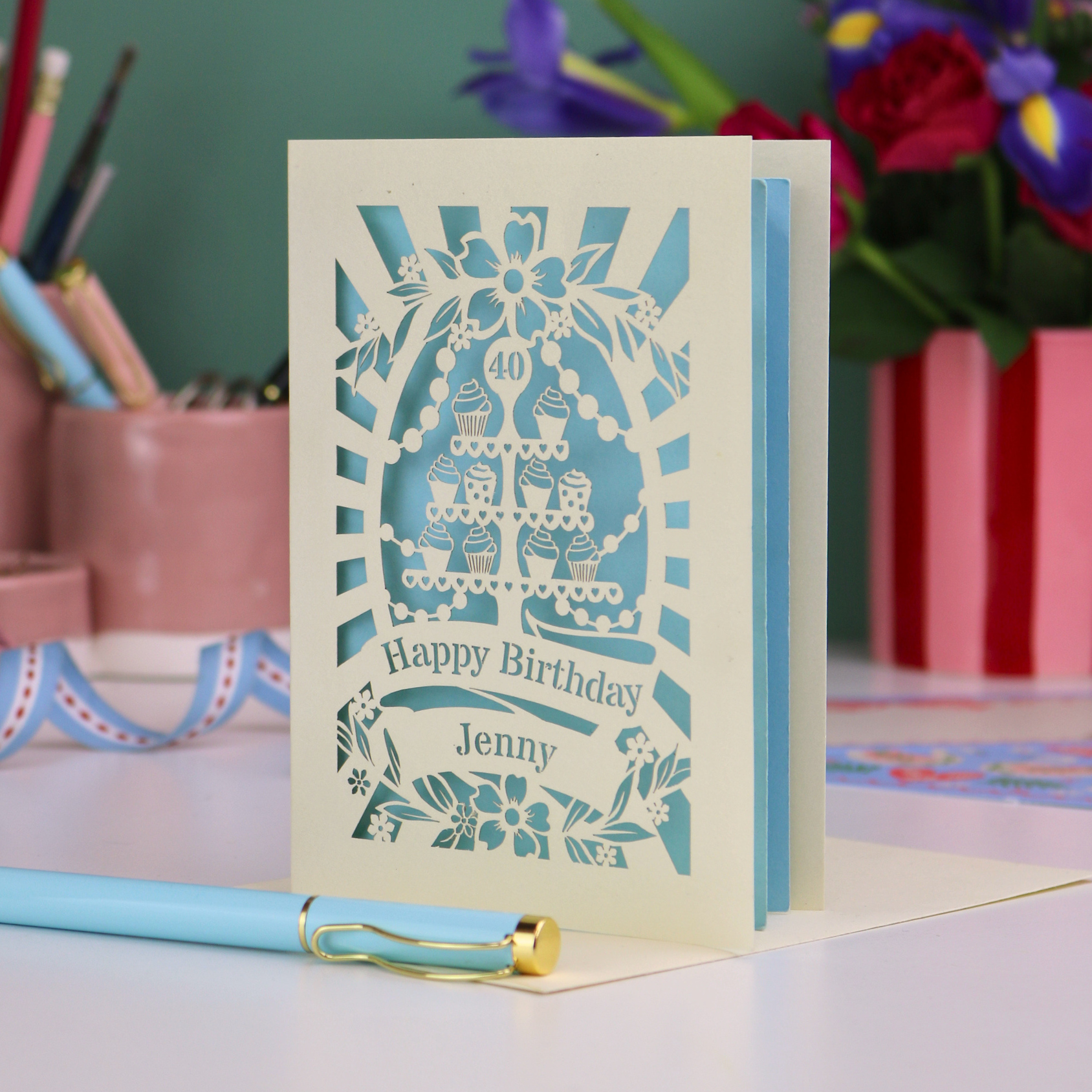 Laser-cut 40th birthday card with decorative cupcake elements and text "Happy Birthday Jenny" on a desk with stationery items.