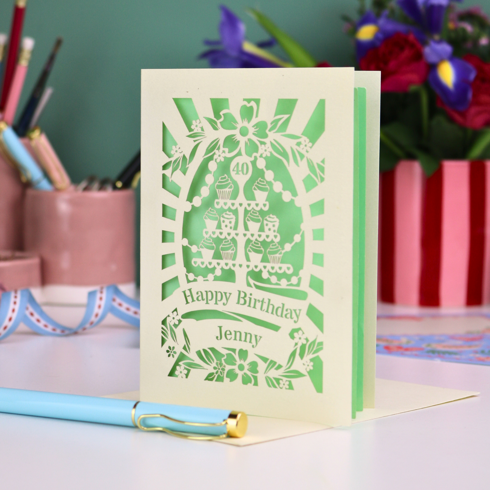 Personalized 40th birthday card with decorative cut-out cupcakes on a cake stand design on a desk with stationery items.