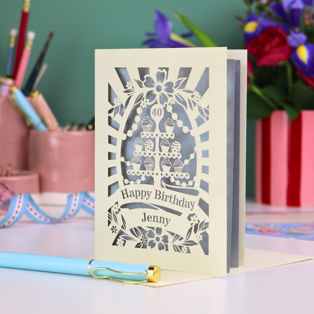 Personalized 40th birthday card with decorative cut-out cupcakes on a cake stand design on a desk with stationery items.
