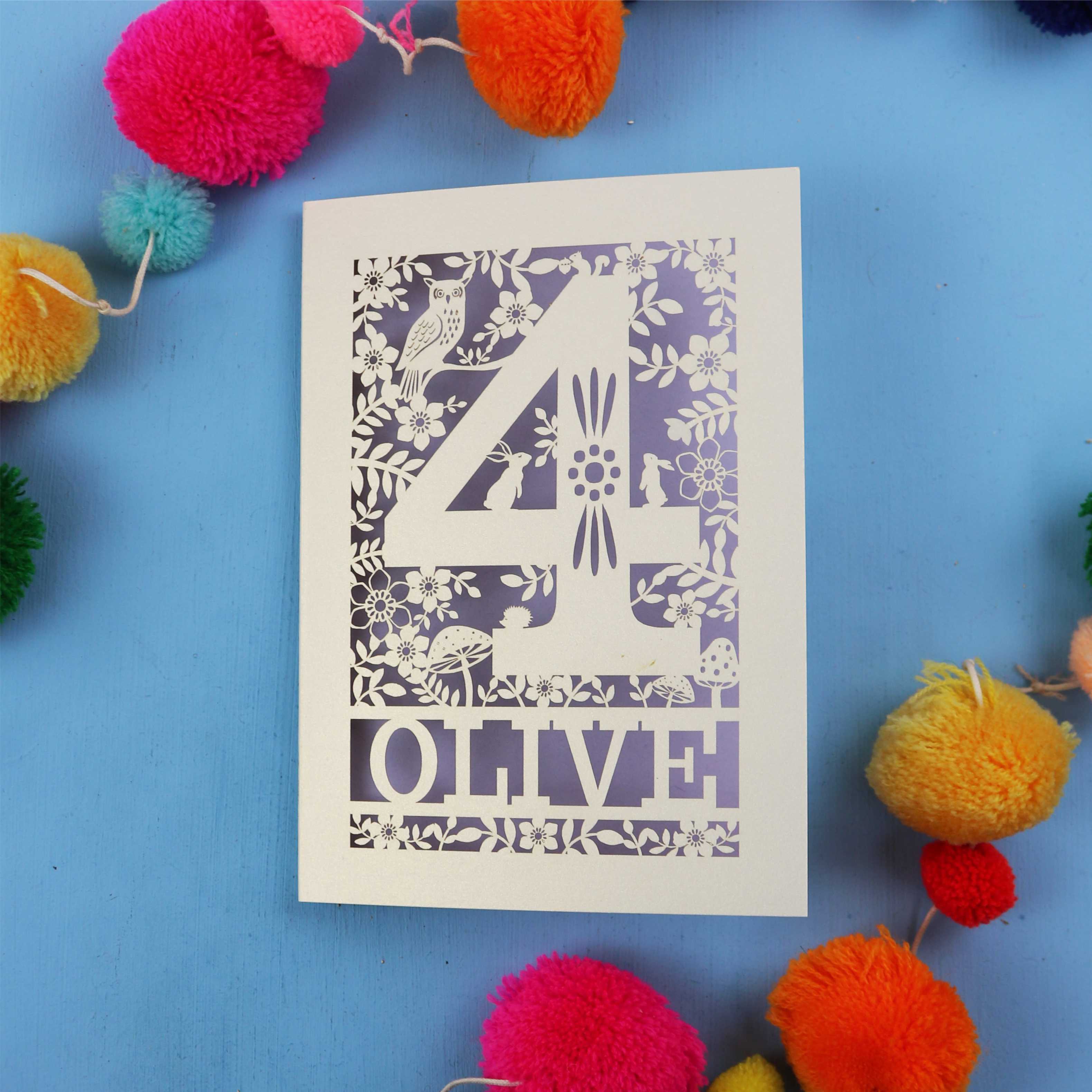 Woodland themed personalised laser cut 4th birthday card with number '4' and 'OLIVE' text on a blue background with colourful pom-poms.