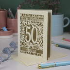 A laser cut 50th birthday card, personalised with the number 50 and the name Annie, on a white desk with stationery items. 