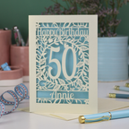A cut out 50th birthday card, personalised with the number 50 and the name Annie, on a white desk with stationery items. 