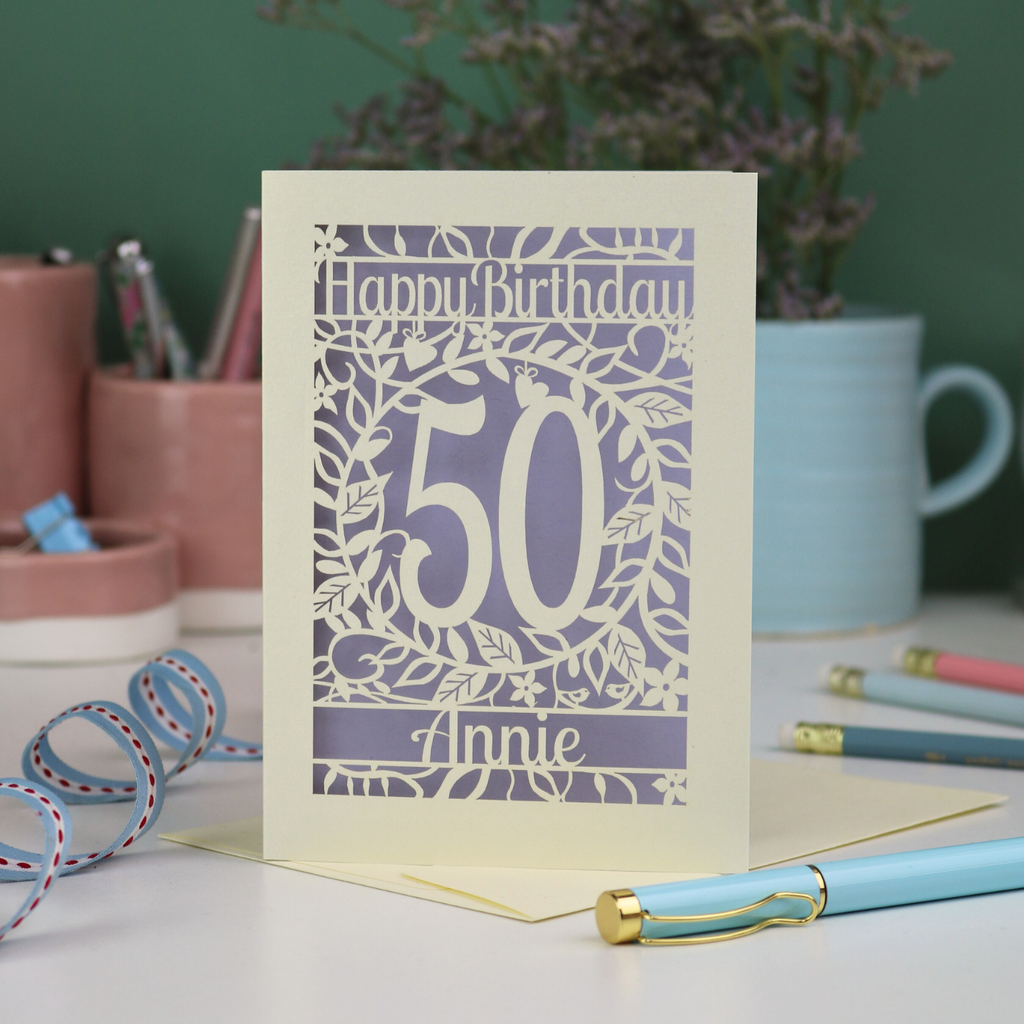 A 50th birthday card, personalised with the number 50 and the name Annie, on a white desk with stationery items. 