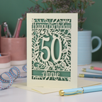 A laser cut keepsake 50th birthday card, personalised with the number 50 and the name Annie, on a white desk with stationery items. 