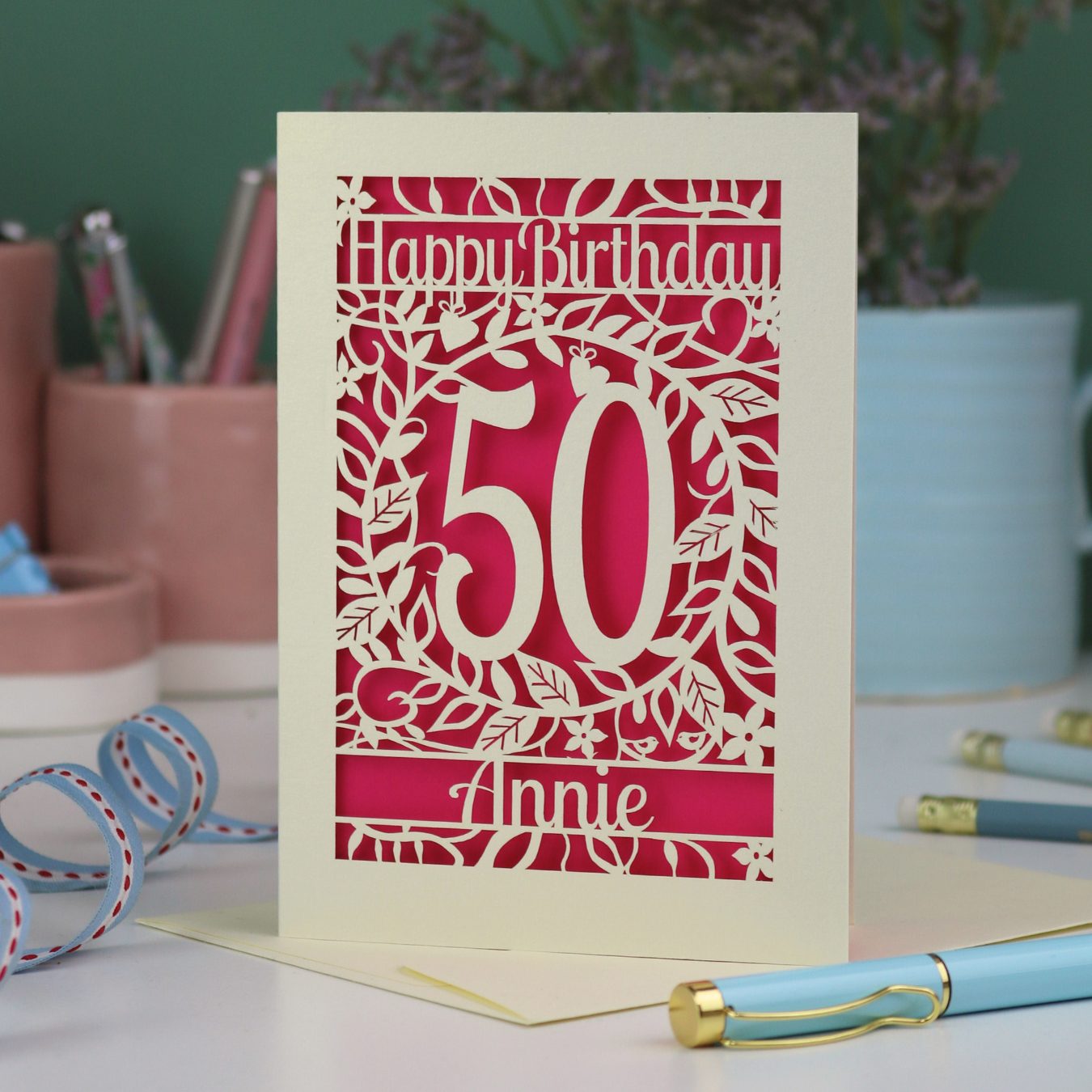 A paper cut 50th birthday card, personalised with the number 50 and the name Annie, on a white desk with stationery items. 