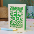A cream 55th anniversary card with green laser-cut floral details, featuring the words 'MAGGIE & RON', the number 55 in the centre and the words  and 'amazing years' underneath. A light blue pen with a gold tip rests in front of the card on a white surface, with a blurred background showing a plant and decorative items.