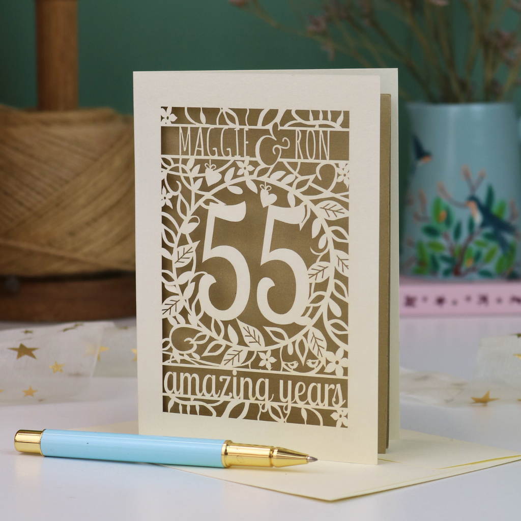 A cream 55th anniversary card with green laser-cut floral details, featuring the words 'MAGGIE & RON', the number 55 in the centre and the words  and 'amazing years' underneath. A light blue pen with a gold tip rests in front of the card on a white surface, with a blurred background showing a plant and decorative items.