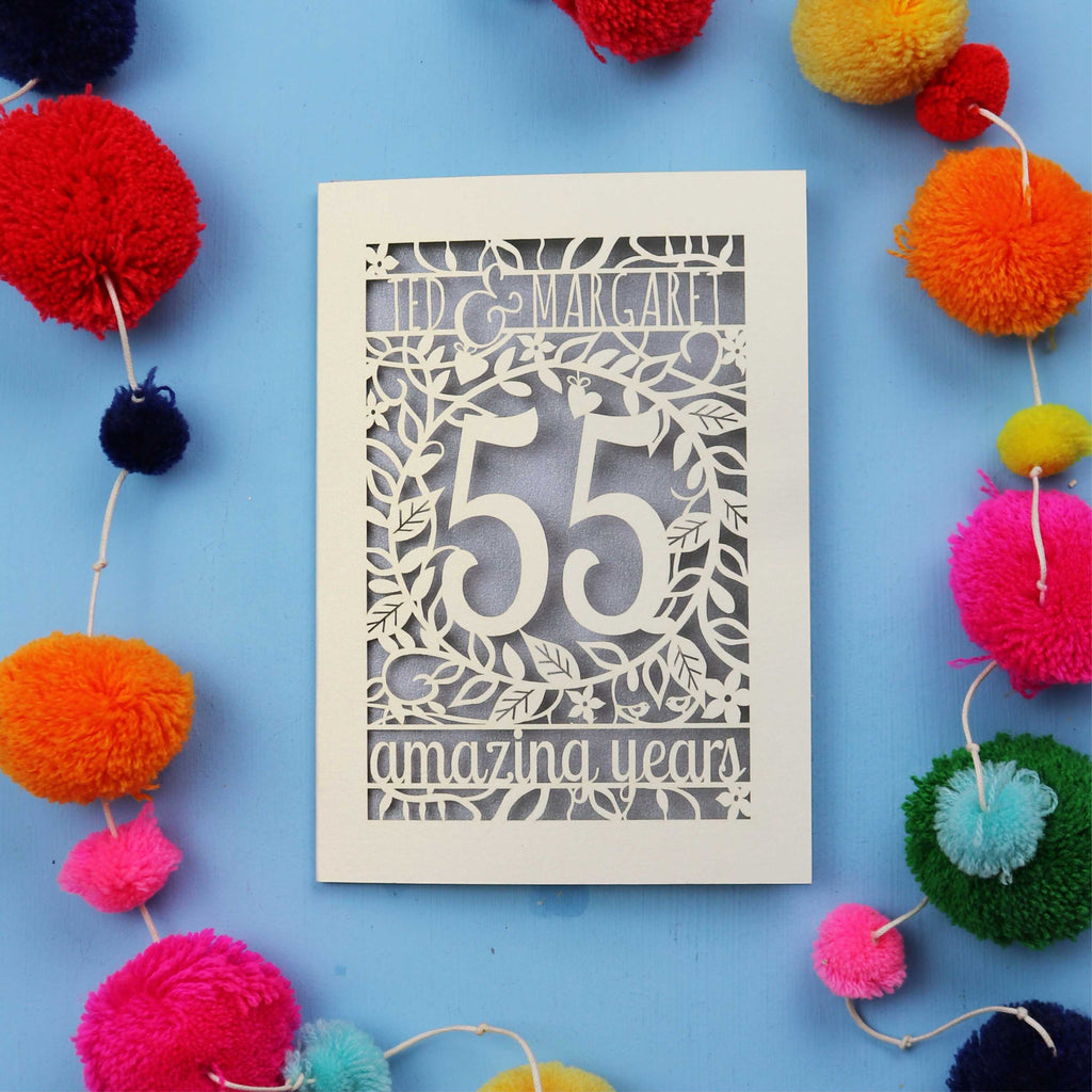 Laser cut Emerald anniversary Card with '55 amazing years' design on a blue background with colourful pom-poms.