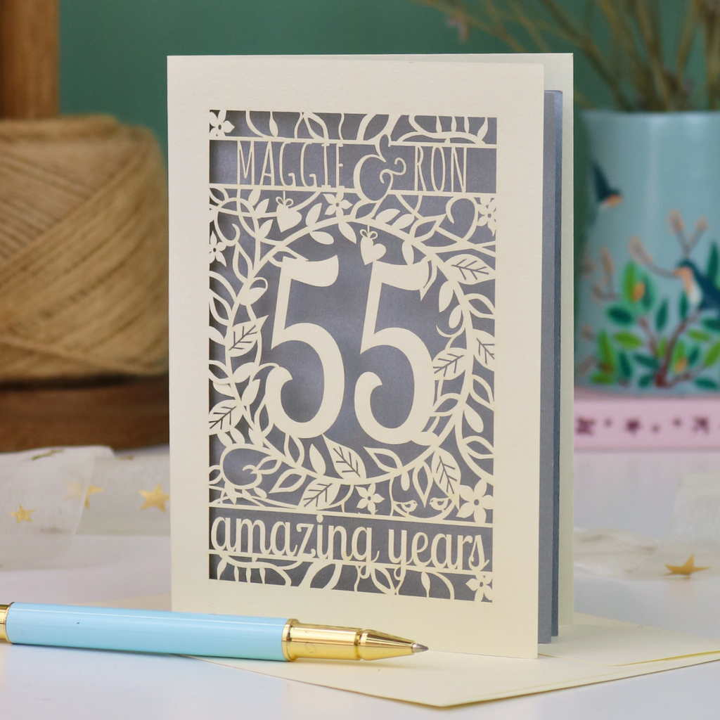 A cream emerald anniversary card with green laser-cut floral details, featuring the words 'MAGGIE & RON', the number 55 in the centre and the words  and 'amazing years' underneath. A light blue pen with a gold tip rests in front of the card on a white surface, with a blurred background showing a plant and decorative items.