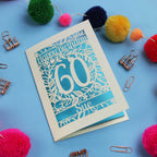 Laser cut 60th Birthday card with 'Happy Birthday 60 Sue' on a blue background with colourful pom-poms and paper clips.