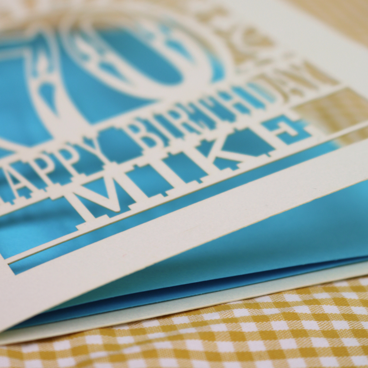 A close-up of a layered paper cut birthday card with laser-cut text reading 'Happy Birthday Mike' in cream over bright blue and gold layers, resting on a yellow-and-white checked fabric surface.