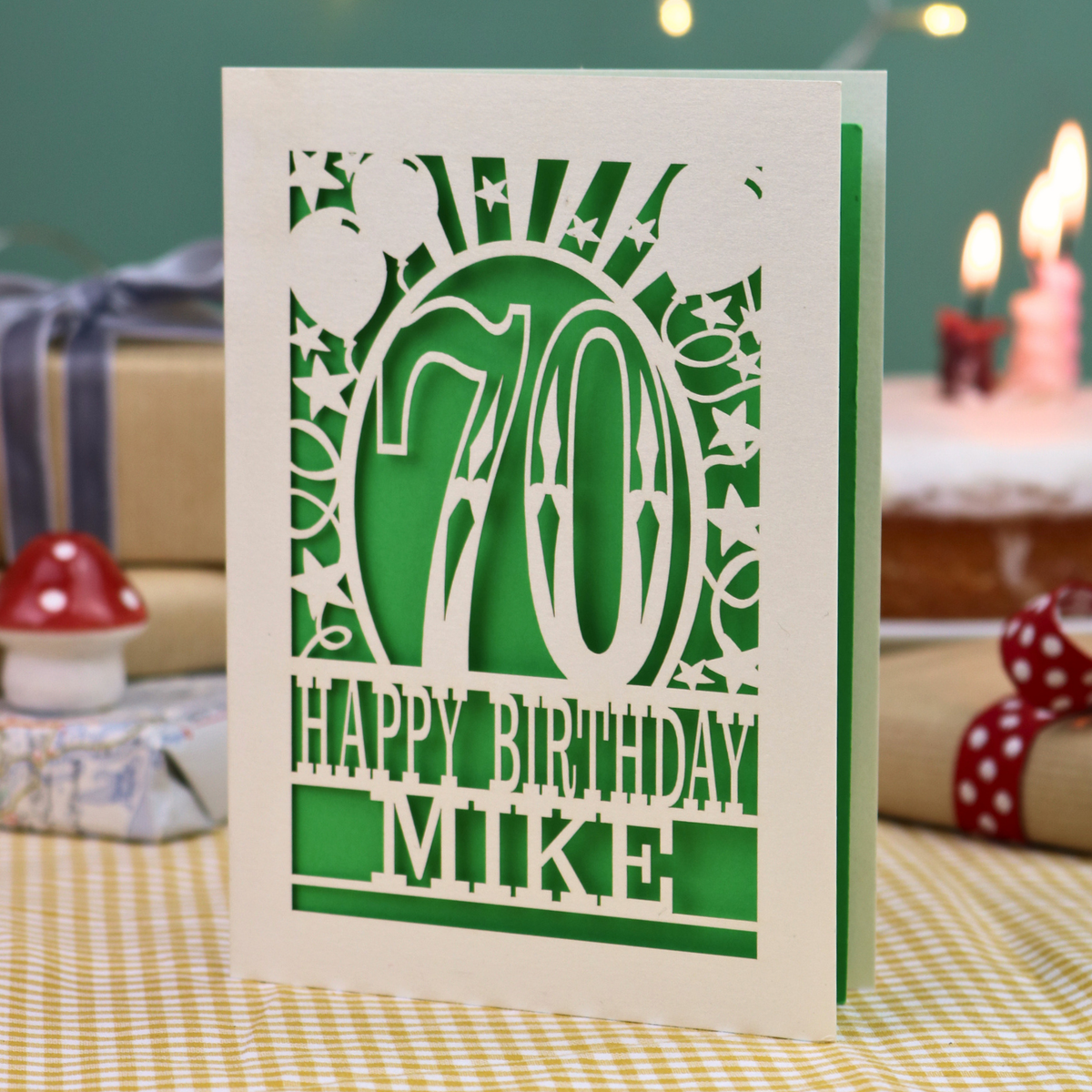 A personalised laser-cut 70th birthday card for a 70th birthday, featuring a cream cut out '70' design with green backing and the text 'HAPPY BIRTHDAY MIKE', displayed on a checked tablecloth with blurred birthday decorations in the background.